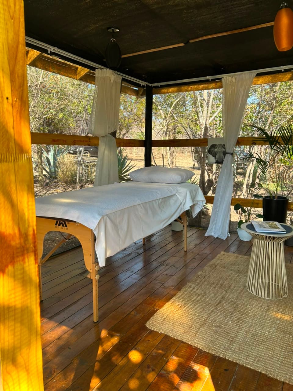 A massage table made up under the open-sided spa sala, with curtains and bush beyond