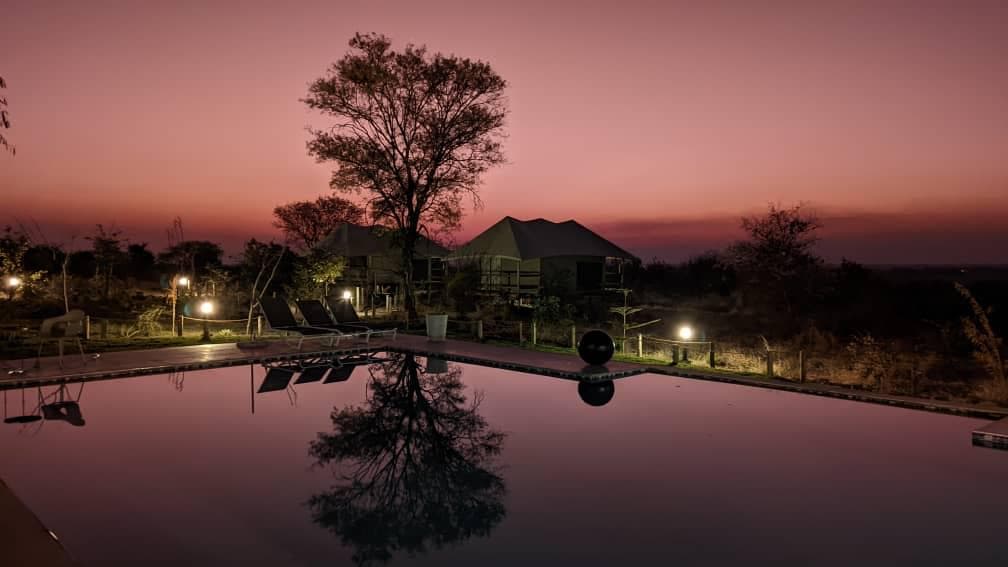 Sunset over the pool at Xhabe Safari Lodge, tented chalets silhouetted against a pink sky and mirrored in the water
